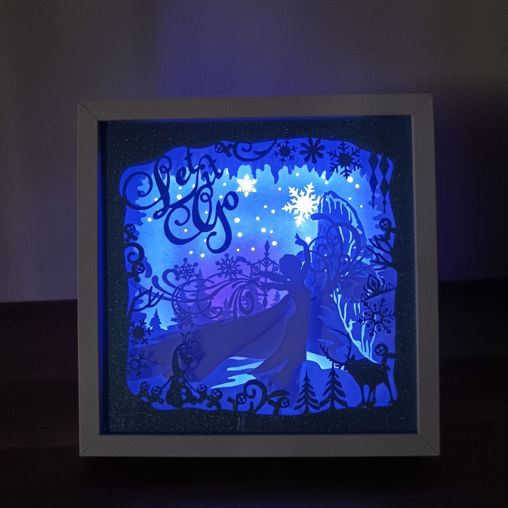 Shadow box lumineuse artisanale inspirée d'un royaume de glace enchanté. Décor multicouche avec reine de glace, flocons scintillants et paysage hivernal illuminé par des LED multicolores - Les Créations des A