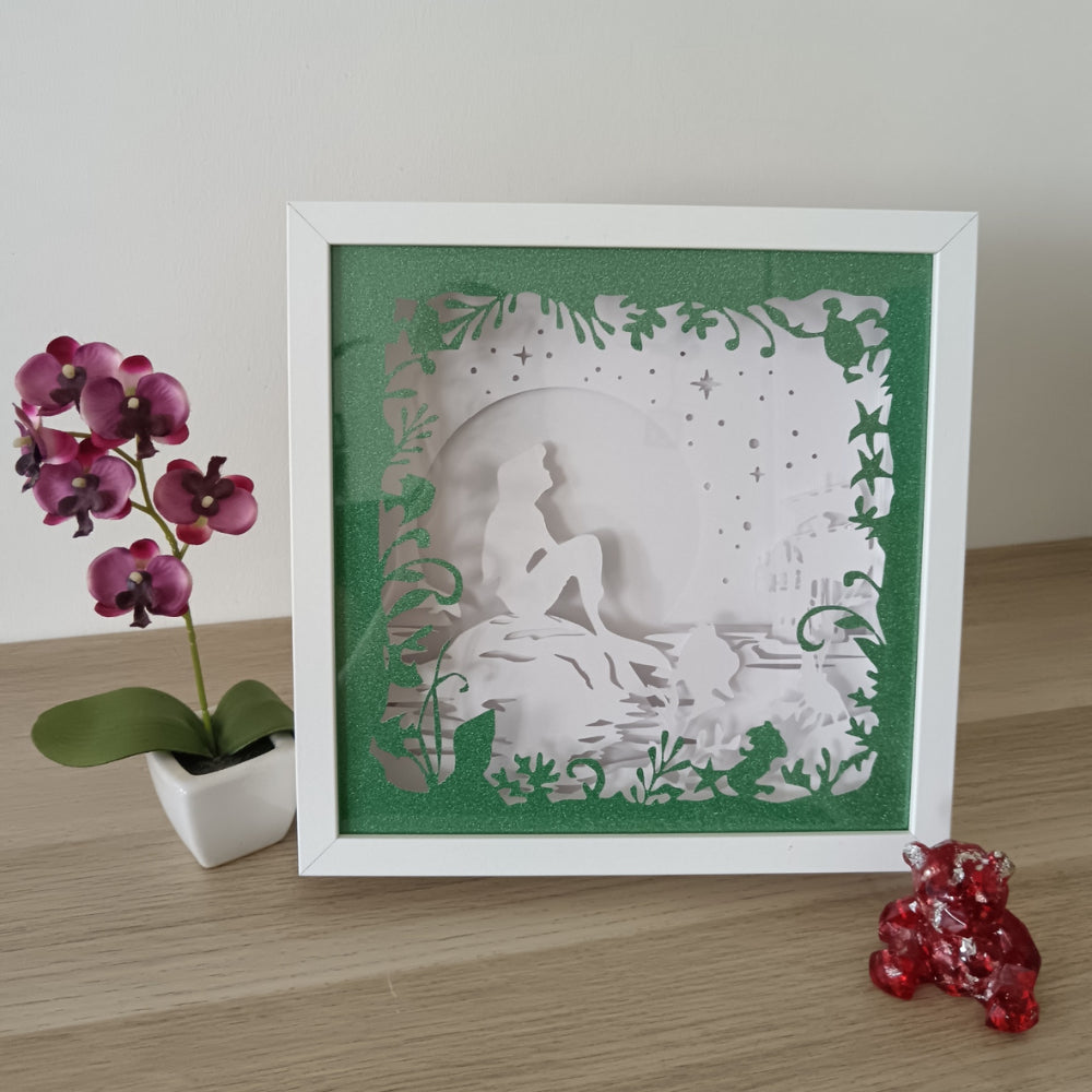 Shadow box lumineuse artisanale représentant une sirène assise au bord de l'eau dans un décor marin féérique. Création multicouche avec éclairage LED et ambiance océanique - Les Créations des A