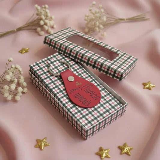 Porte-clé en cuir rouge personnalisé gravé, présenté sur sa boîte cadeau à carreaux, décoration étoiles dorées et fleurs séchées fond rose - Les Créations des A