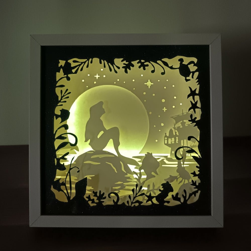Shadow box lumineuse artisanale représentant une sirène assise au bord de l'eau dans un décor marin féérique. Création multicouche avec éclairage LED et ambiance océanique - Les Créations des A