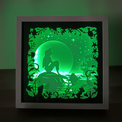 Shadow box lumineuse artisanale représentant une sirène assise au bord de l'eau dans un décor marin féérique. Création multicouche avec éclairage LED et ambiance océanique - Les Créations des A
