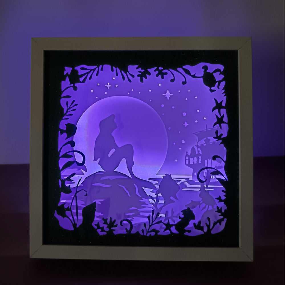 Shadow box lumineuse artisanale représentant une sirène assise au bord de l'eau dans un décor marin féérique. Création multicouche avec éclairage LED et ambiance océanique - Les Créations des A
