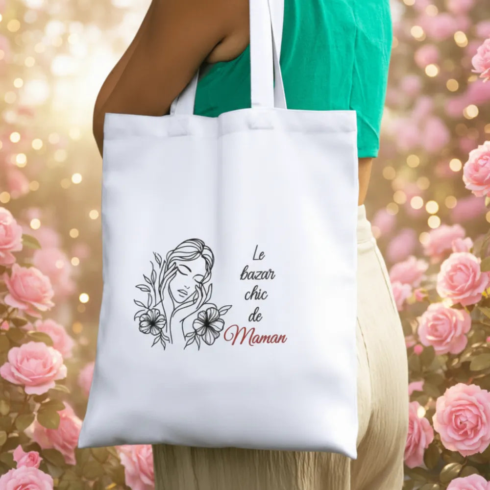 Femme portant un tote bag blanc "Le bazar chic de Maman" dans un jardin fleuri – cadeau maman - Les Créations des A