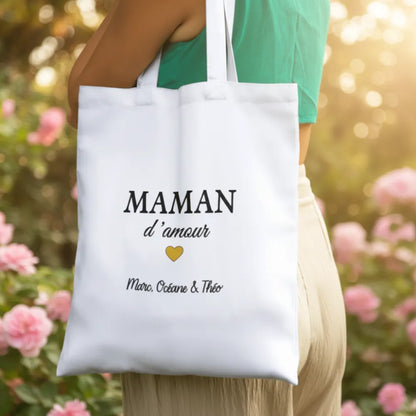 Femme portant un tote bag blanc "Maman d'amour" dans un jardin fleuri – cadeau fête des mères - Les Créations des A