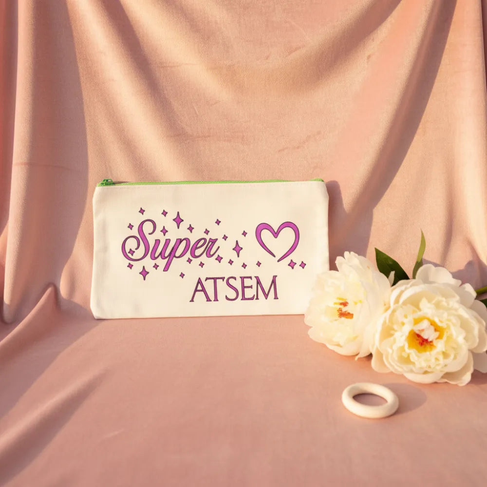 Trousse blanche "Super ATSEM" avec cœur violet et étoiles, posée sur velours rose avec pivoines blanches – Cadeau fin d'année ATSEM – Les Créations des A