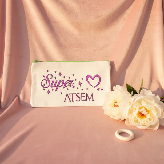 Trousse blanche "Super ATSEM" avec cœur violet et étoiles, posée sur velours rose avec pivoines blanches – Cadeau fin d'année ATSEM – Les Créations des A